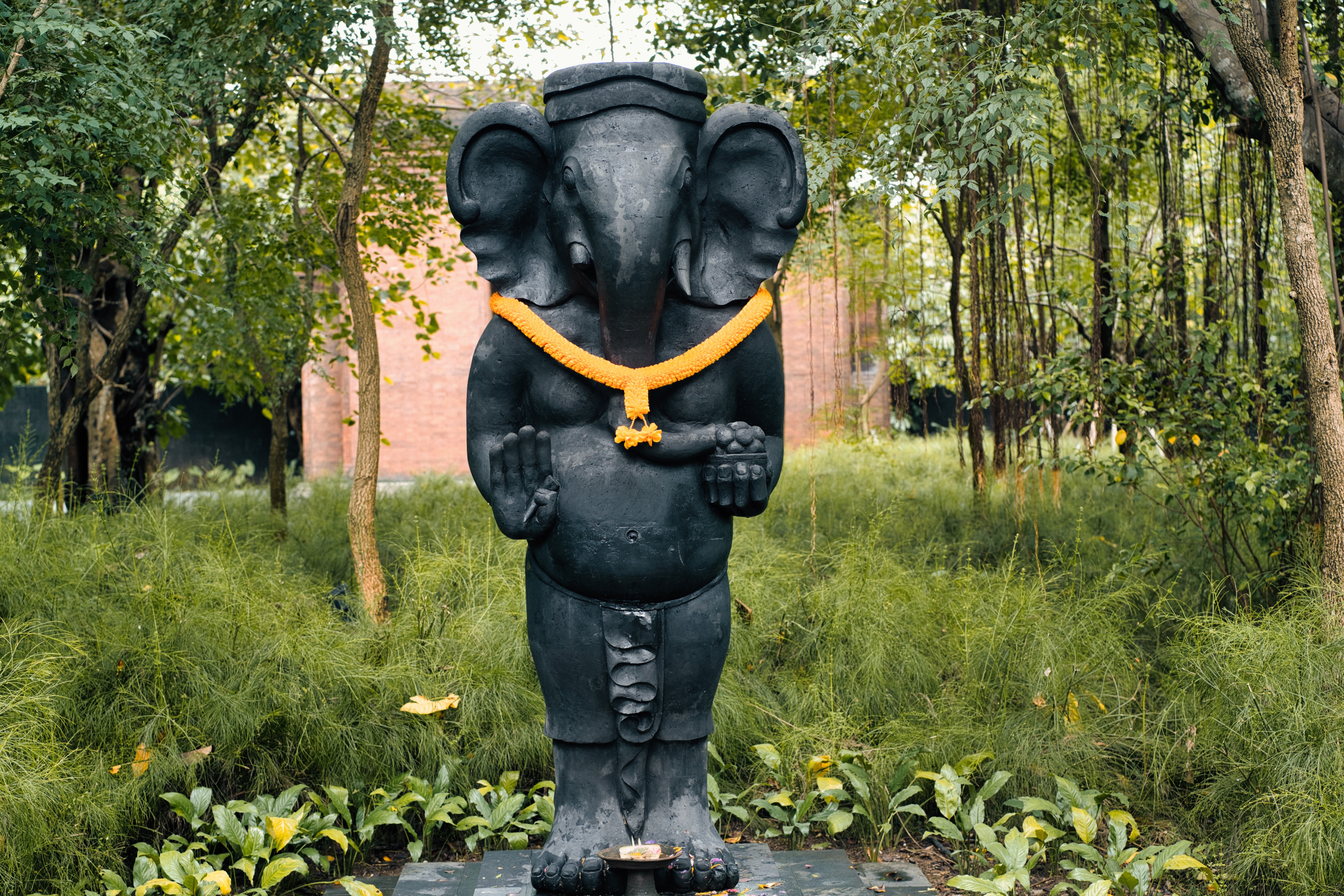 A large dark three-headed statue draped in a marigold garland, in a temple garden